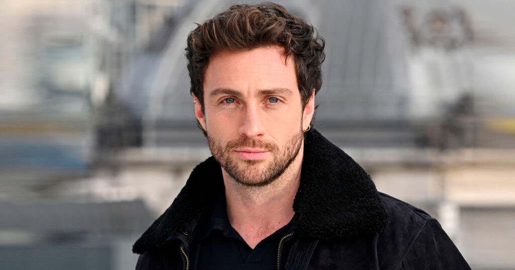 Retrato do ator Aaron Taylor-Johnson, que veste uma jaqueta escura e olha para a câmera em um ambiente externo.