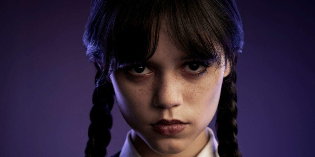 Close-up do rosto de Jenna Ortega como Wandinha Addams, com suas tranças características e olhar sério, contra um fundo roxo.