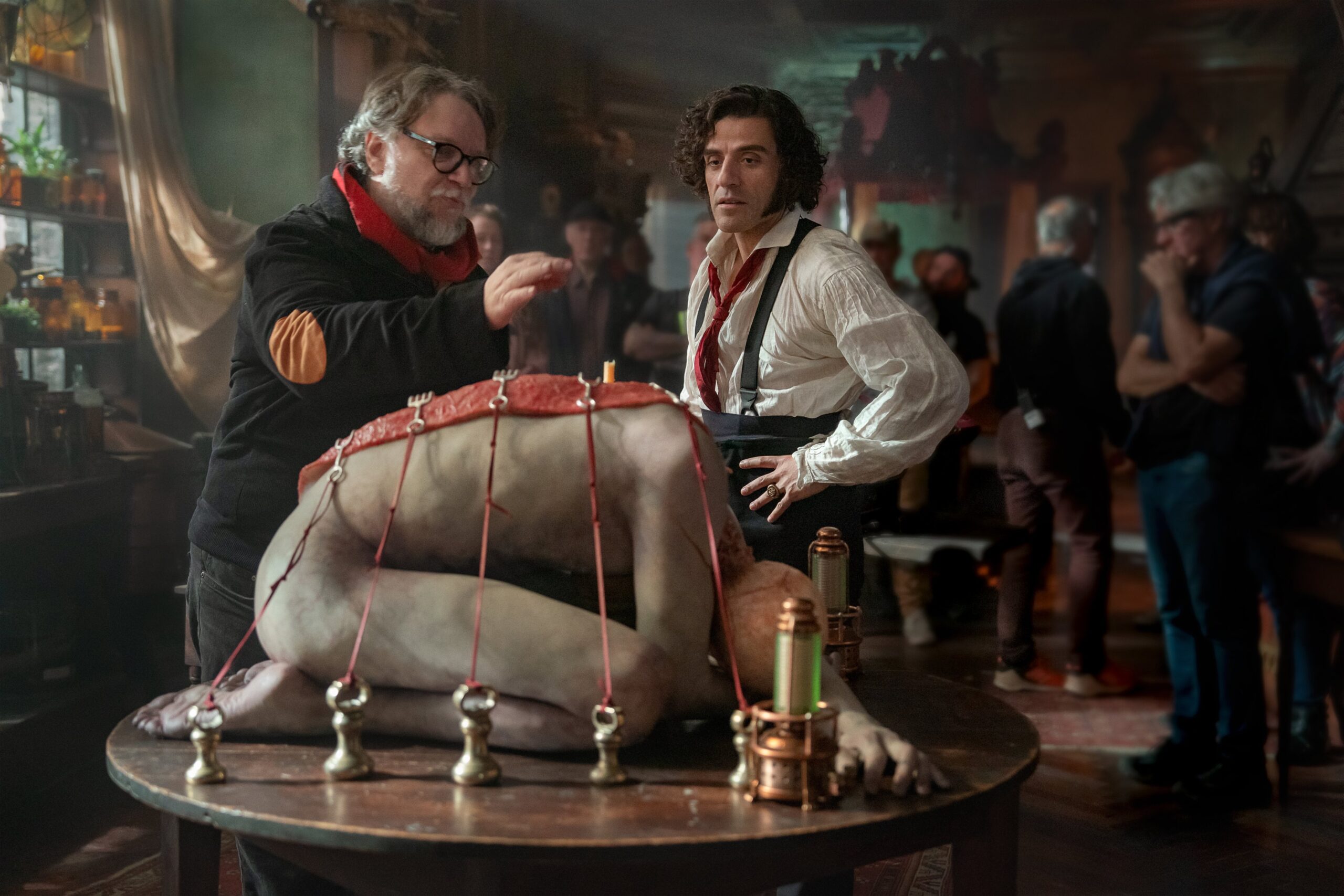 O diretor Guillermo del Toro e o ator Oscar Isaac nos bastidores do filme Frankenstein, em frente a uma mesa de laboratório onde está o protótipo da criatura.
