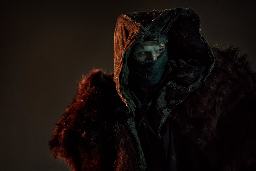 O ator Jacob Elordi com a maquiagem e figurino de O Monstro no filme Frankenstein de Guillermo del Toro. Ele usa um capuz e roupas de pele, com um olhar sombrio.