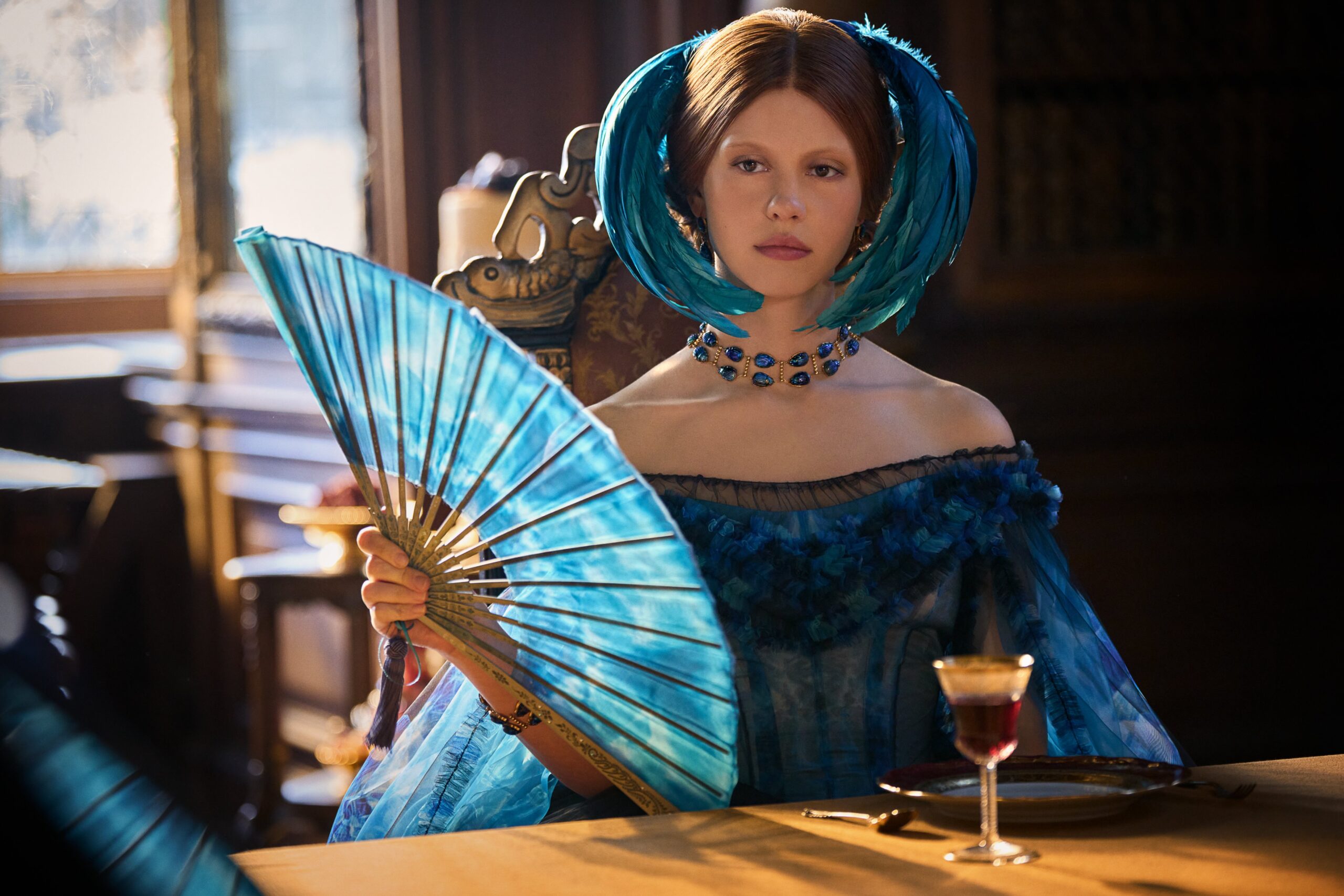 A atriz Mia Goth em sua caracterização como a personagem Elizabeth no filme Frankenstein. Ela está sentada à mesa com um leque azul e um vestido da mesma cor, em um cenário de época.